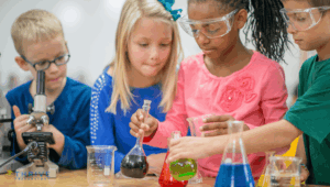 four children are working with beakers that have blue, green, red, and dark green, yellow and clear liquids. The table also has a microscope. the children are standing in the following order: little boy withbloind haiar and blue eyeglasses and long sleeveld tshirt, littel girl about 9 years old with blond hair and a blue ribbon in her hair, wearing a blue shirt with sequins accents on the sleeve, next to a little girl, about 9 years old, with braids, a pink long--sleeved tshirt that has a rose on the top left hand side of it is holding a beaker with dark liquid and about to pour it into a smaller beaker. she has on goggles. the lastboy is earing a green tshirt, hasa brown hair and is wearing safety goggles. he is holding a beaker with light freen liquid and about to pour it into the beaker of red liquid.